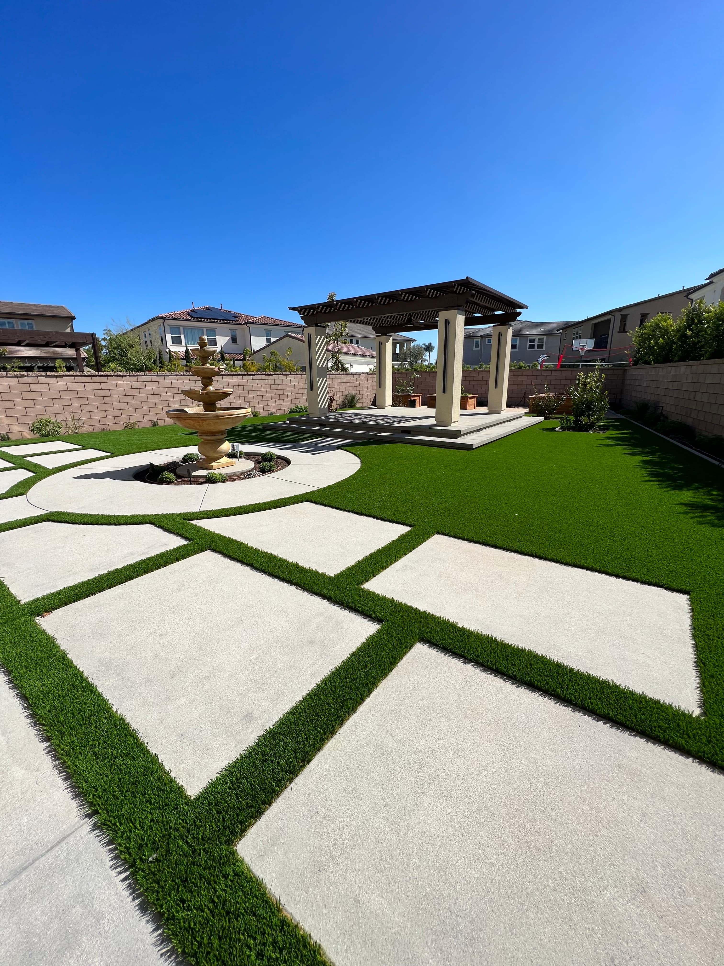 Modern backyard with a tiered stone fountain, concrete pavers, artificial turf, and a wooden pergola.