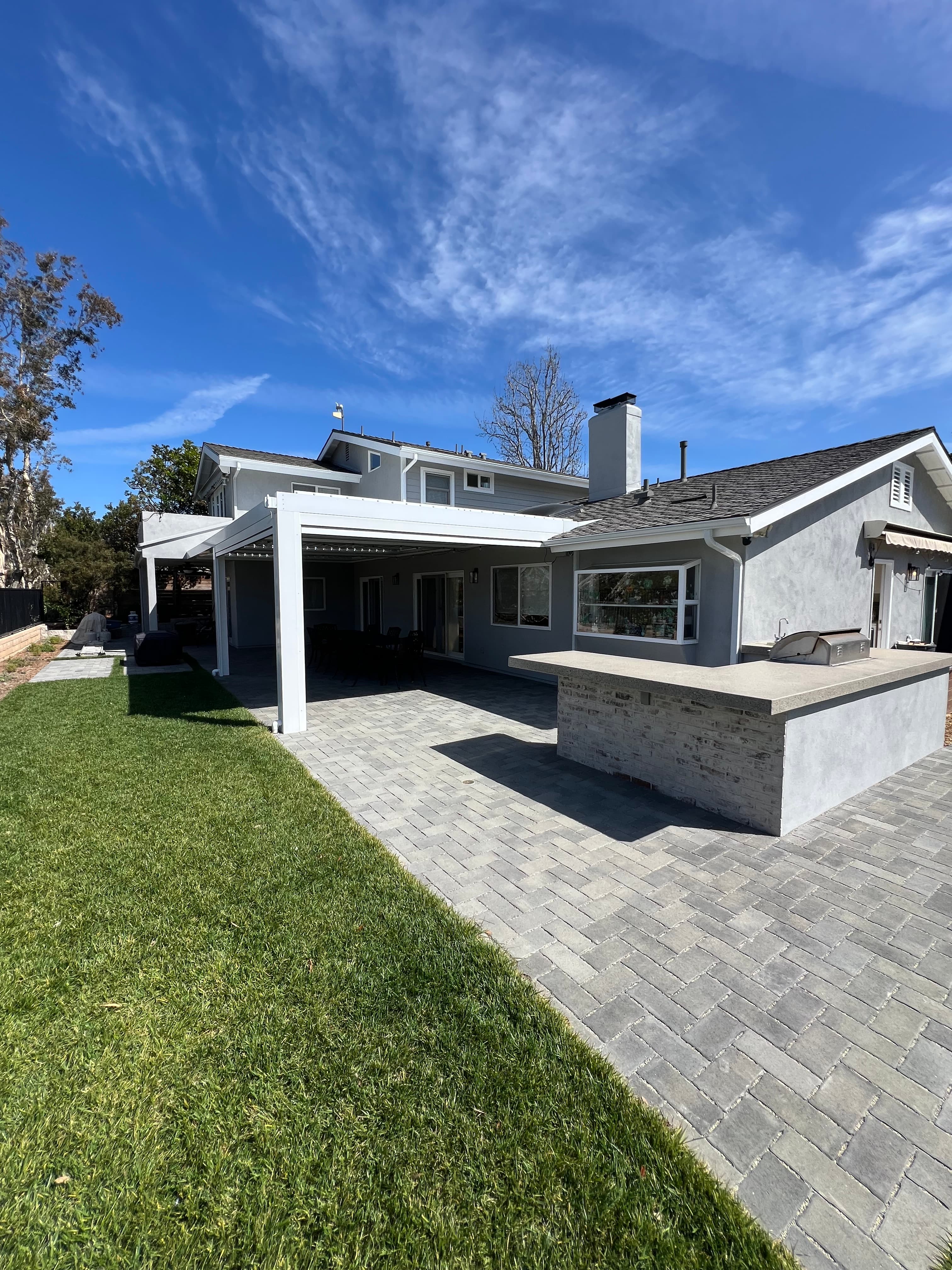Modern backyard with a gray stone patio, white pergola, outdoor kitchen, and green lawn.