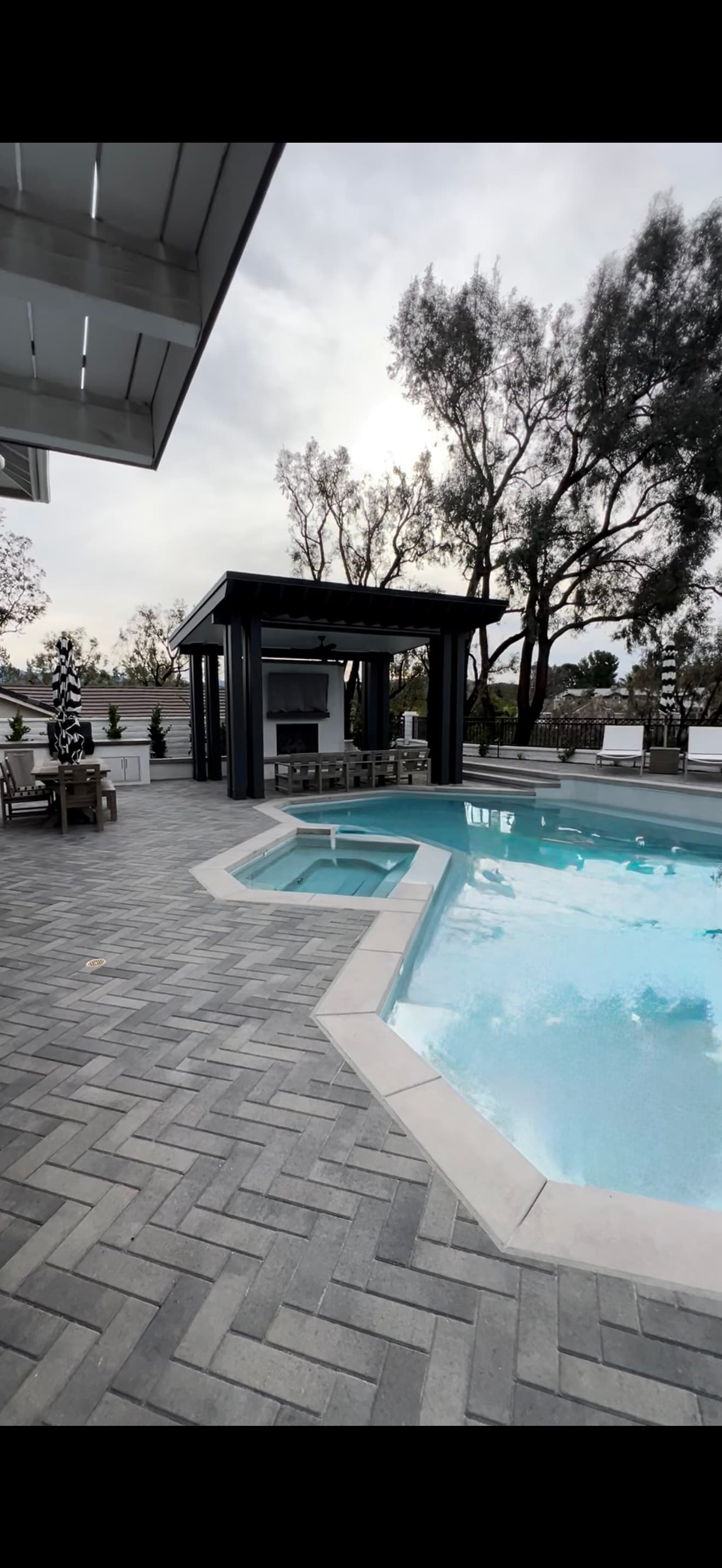 Modern swimming pool and spa surrounded by gray herringbone pavers and a black outdoor pavilion.