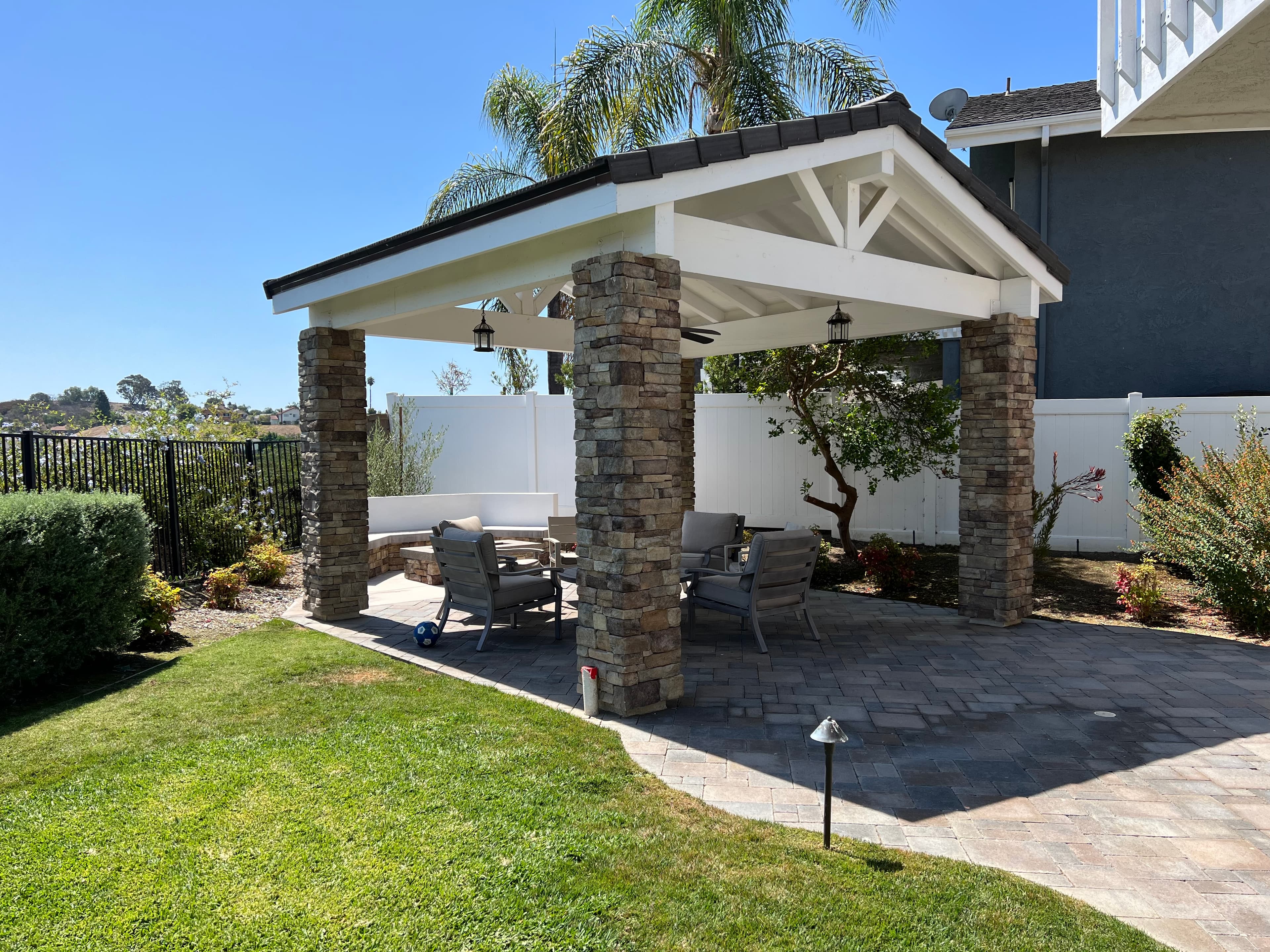 White gabled pavilion with stone pillars over a paved patio with grey outdoor seating.
